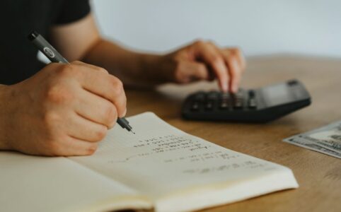 Person's hands using a calculator over house blueprints and financial documents on a bright, modern desk, representing the...